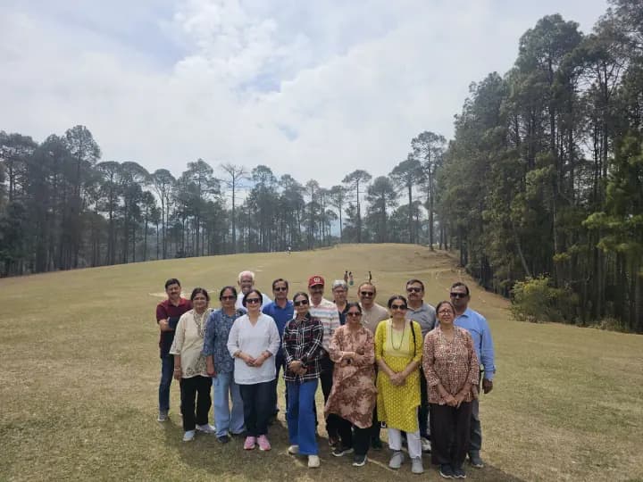 Large family group gathered across a wide alpine meadow bordered by Himalayan pines